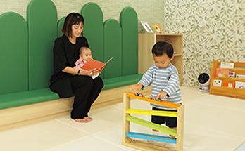 プレイルームで赤ちゃんに絵本の読み聞かせをする保護者とおもちゃで遊ぶこども
