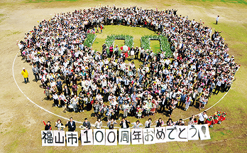 記念写真：福山市100周年おめでとう！と書かれた紙を掲げる人たちと植物で書いた大きな「100」の地上文字を円状に囲っている人々が写っている。